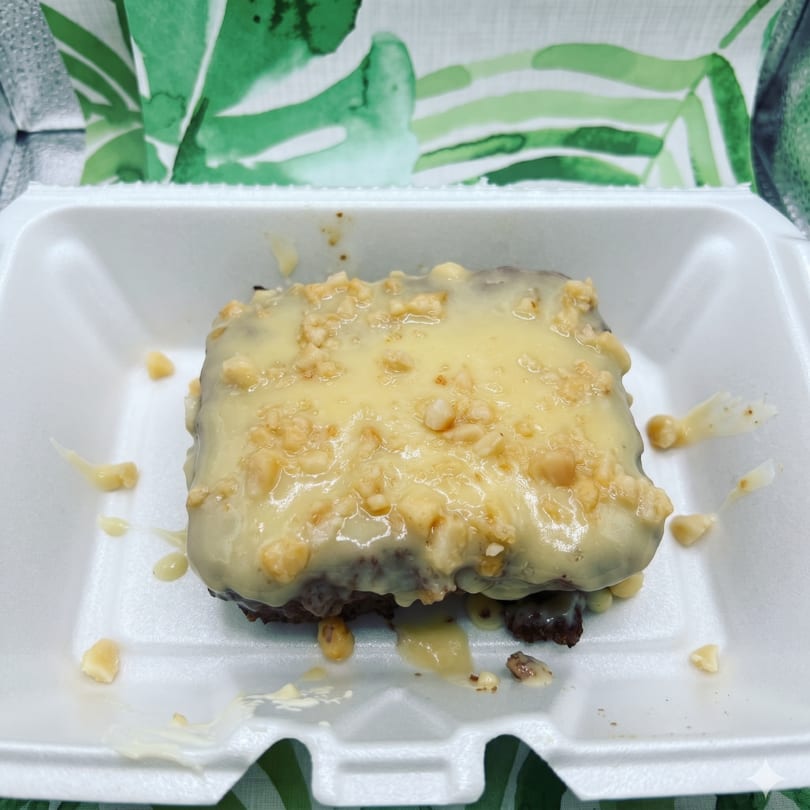 A chocolate brownie with creamy white frosting and chopped nuts in a white takeout container.