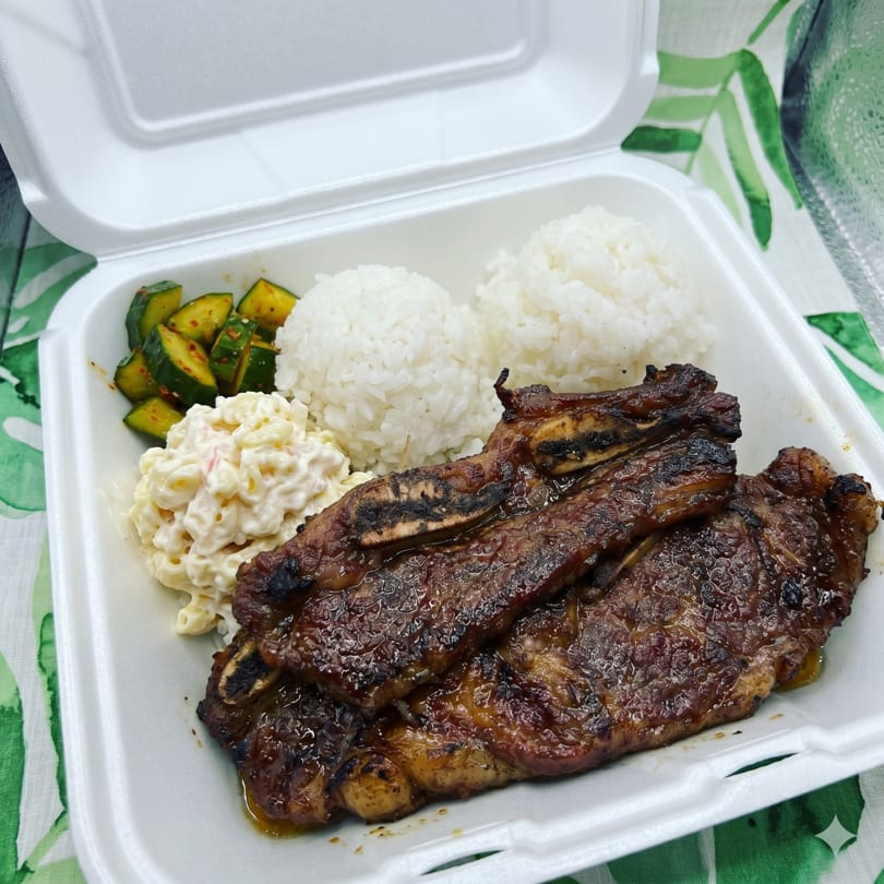 Grilled short ribs, two scoops of white rice, macaroni salad, and cucumber salad in a takeout container.